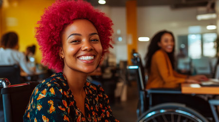 A happy black African American woman in a wheelchair with vibrant pink afro hair smiles warmly in a lively, inclusive office setting.の素材