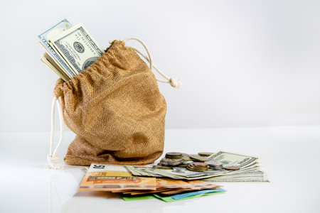 A brown burlap sack filled with US dollar bills sits on a white table next to scattered coins and bills.の写真素材