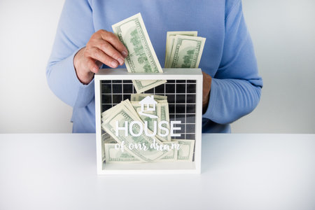 A person puts money into a clear box with a House of Our Dreams sign, symbolizing saving for a house.の写真素材
