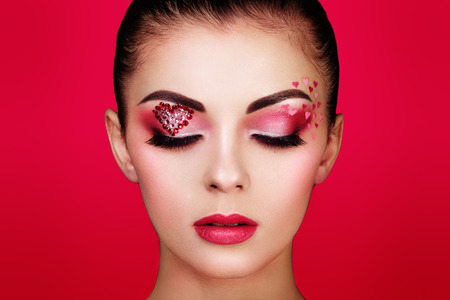 Face of beautiful woman with holiday makeup heart. Valentine's day make-up. Lips in pink lipstick. Makeup detail. Face of girl on a red backgroundの写真素材