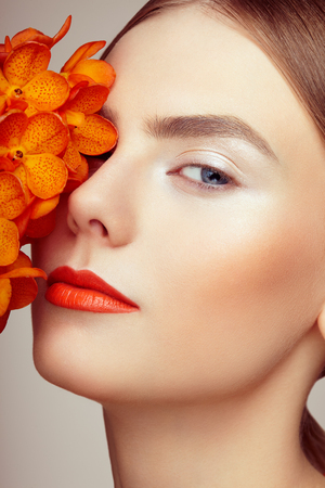 Portrait of beautiful young woman with orchid. Brunette woman with luxury makeup. Perfect skin. Eyelashes. Cosmetic eyeshadow. Orange flowersの写真素材