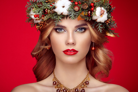 Portrait of beautiful young woman with Christmas wreath. Beautiful New Year and Christmas tree holiday hairstyle and makeup. Beauty girl portrait isolated on red background. Colorful makeup and hairの写真素材