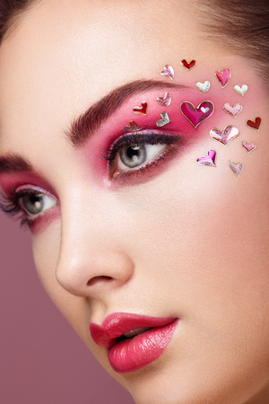 Face of Beautiful Woman with Holiday Makeup Heart. Valentine's Day Make-up. Lips in Pink Lipstick. Makeup detail. Face of Girl on a Pink Backgroundの写真素材