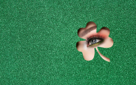 Conceptual photo of St. Patrick's Day. The Eye of the Young Beautiful Woman with Bright Golden Shadows and Expressive Eyebrows, Looks in the Shamrock Leaves Pattern out of Green Paperの写真素材