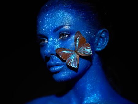 Fashion model woman in blue bright sparkles and neon lights posing in studio.  Portrait of beautiful sexy woman with blue butterfly. Art design colorful glitter glowing make upの写真素材
