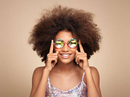 Beauty portrait of African American girl in colored holographic sunglasses. Beautiful black woman on pink background. Cosmetics, makeup and fashionの写真素材