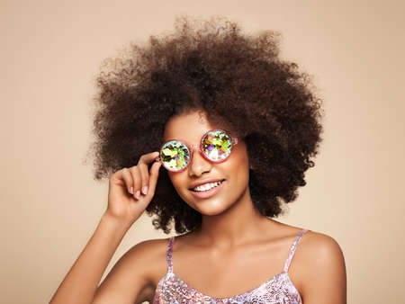 Beauty portrait of African American girl in colored holographic sunglasses. Beautiful black woman on pink background. Cosmetics, makeup and fashionの写真素材