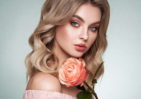 A beautiful young woman with shiny wavy blonde hair. Model with healthy skin, close up portrait. Girl with a rose flowerの写真素材
