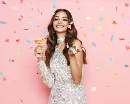 Beautiful woman in cocktail dress posing with glass of champagne under confetti. Model with curly hair. Christmas or Happy New Year. Holiday and Partyの写真素材