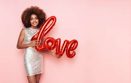 A beautiful young African-American woman with a balloon in the shape of the inscription love on a pink background. Woman on Valentine's Day. Symbol of loveの写真素材