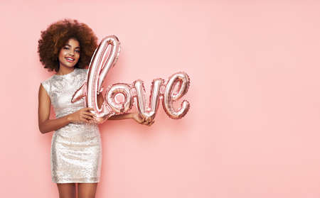 A beautiful young African-American woman with a balloon in the shape of the inscription love on a pink background. Woman on Valentine's Day. Symbol of loveの写真素材