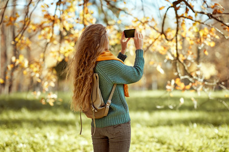 A young red-haired woman walks through the autumn park. A girl with a smartphone takes photos in an autumn forestの写真素材