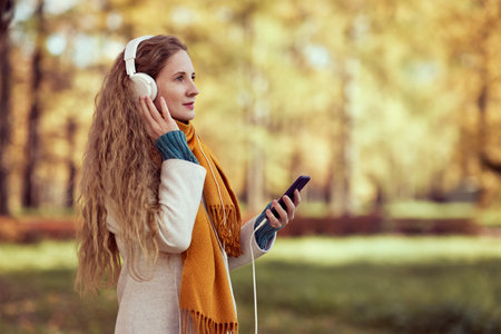 A young red-haired woman in a white coat walks in an autumn park. A girl listens to music in the autumn forestの写真素材