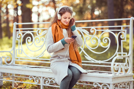 A young red-haired woman is sitting on a bench in an autumn park. A girl listens to music in the autumn forestの写真素材