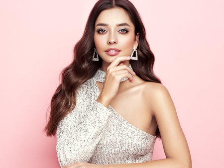 Beautiful woman in cocktail dress posing on pink background. Beauty model with long curly hair. Christmas or New Year festivities. Jewelry and manicureの写真素材