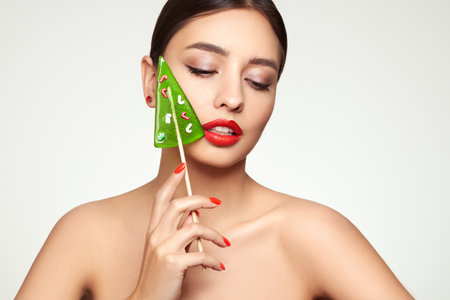 Beauty model girl eating Christmas tree shape lollipop.  Beautiful young woman with clean fresh skin. Girl beauty face care. Facial treatment. Cosmetology, beauty and spa. Red nails manicureの写真素材