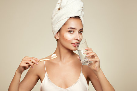 Image of a young happy positive woman in towel and with cosmetic patches on the eyes, brushing her teeth isolated over beige wall backgroundの写真素材