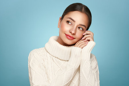 Portrait of young happy beautiful woman smiling and looking away, isolated on blue background in a warm white sweater. Concept of winter promo saleの写真素材