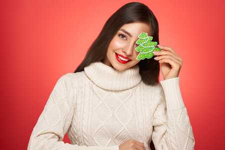 Woman in a white knitted sweater with cookie Christmas tree isolated on red background. Christmas or New Year festivities. Holiday and Partyの写真素材