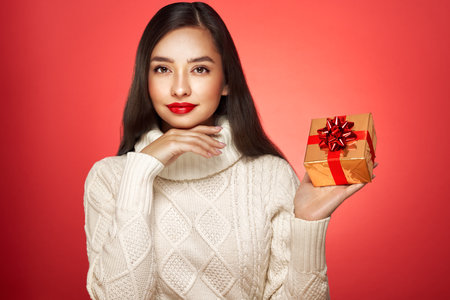 Beautiful in a white knitted sweater with gift box isolated on red background. Christmas or New Year festivities. Holiday and Partyの写真素材