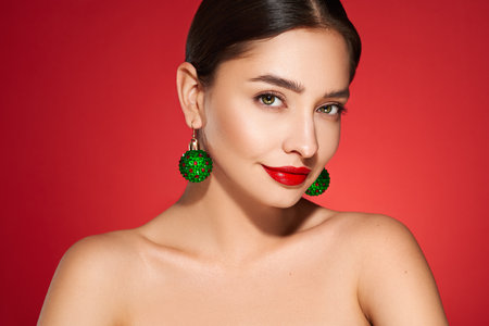 Portrait of beautiful young woman on red background, red lipstick and festive makeup, Christmas toy earrings, shiny glowing skin, Winter holidays conceptの写真素材