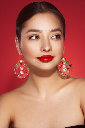 Portrait of beautiful young woman on red background, red lipstick and festive makeup, Christmas toy earrings, shiny glowing skin, Winter holidays conceptの写真素材