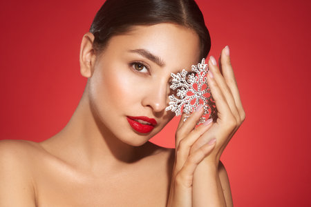 Beautiful young woman with red lipstick, shiny clean skin and face, showing glowing snowflake, near face and smiling, isolated on a red background. Christmas holidays conceptの写真素材