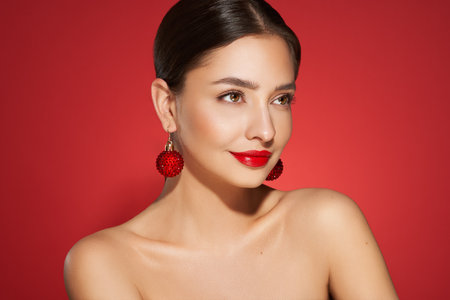 Portrait of beautiful young woman on red background, red lipstick and festive makeup, Christmas toy earrings, shiny glowing skin, Winter holidays conceptの写真素材