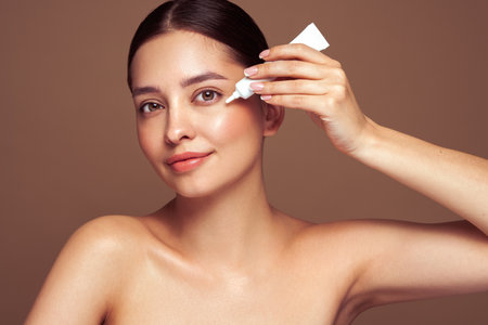 Beautiful woman using cream around the eyes. Video of woman with perfect makeup on beige background. Beauty and skin care conceptの写真素材
