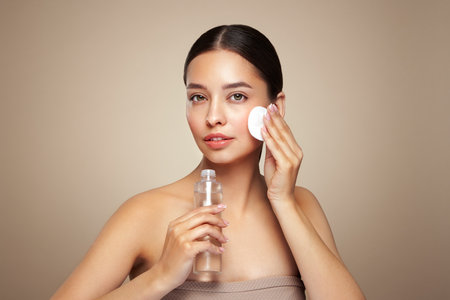 Smiling woman holds bottle with cosmetic tonic. Photo of attractive woman with perfect makeup on beige background. Beauty and skin care concept. Removing makeupの写真素材