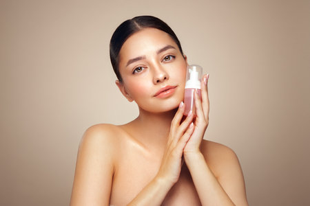 A young woman shows off a bottle with a foam dispenser for washing. Daily skin care cosmetics advertisement. Studio shoot Beige backgroundの写真素材