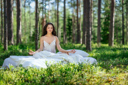 Young woman is resting on a mattress in the lotus position, in summer forest. Girl is sleeping in the woodsの写真素材