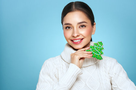 Woman in a white knitted sweater with cookie Christmas tree isolated on blue background. Christmas or New Year festivities. Holiday and Partyの写真素材