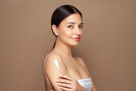 Cropped shot of a young tanned woman moisturizing her shoulder with her hand using a cream isolated on a beige background. Skin care. A white thick spear of cream on the female bodyの写真素材