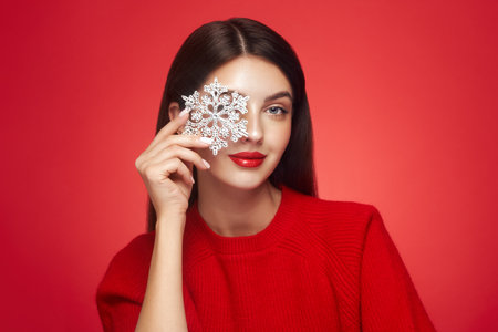 Beautiful young woman with red lipstick, shiny clean skin and face, showing glowing snowflake, near face and smiling, isolated on a red background. Christmas holidays conceptの写真素材