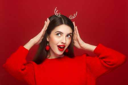Portrait of beautiful young woman on red background, red lipstick and festive makeup, Christmas toy earrings, shiny glowing skin, Winter holidays conceptの写真素材