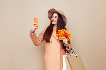 A stylish woman, dressed in fashionable autumn outfit including beige coat and hat, smiles while looking at her smartphone, holding several shopping bags against beige backgroundの写真素材