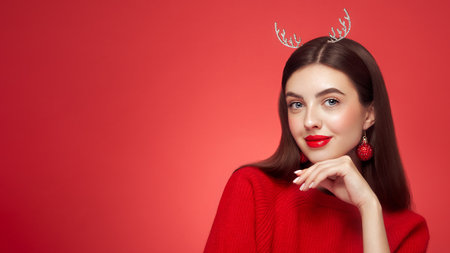 Portrait of beautiful young woman on red background, red lipstick and festive makeup, Christmas toy earrings, shiny glowing skin, Winter holidays conceptの写真素材