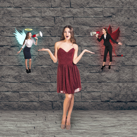 Pretty young woman in red dress thinks, an angel and a devil on grey brick backgroundの写真素材