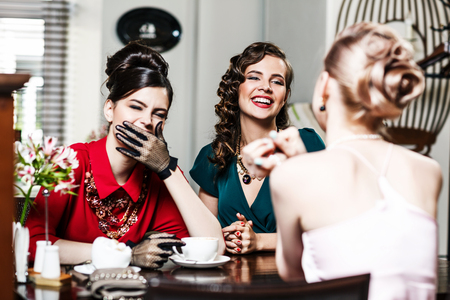 Stylish smiling women talking and drinking coffee sitting inside in cafeの写真素材