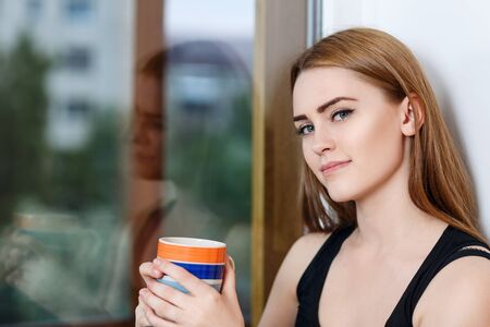 Young woman sitting with cup at windowsill at home in rainy day.の写真素材