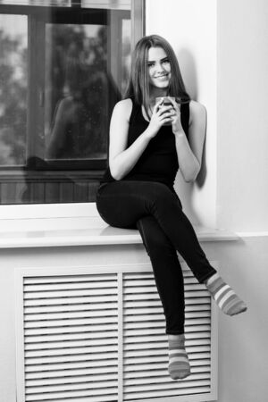 Young woman sitting with cup at windowsill at home in rainy day.の写真素材