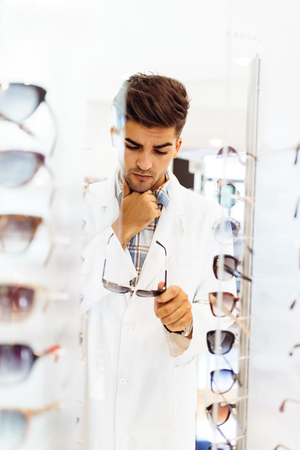 Handsome young doctor choosing eyeglasses frame in optical store.の写真素材
