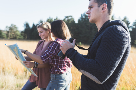 Young friends enjoying outdoor. Two girls standing in deep grass, smiling and looking at map to find best route while young man looking through binoculars.の写真素材