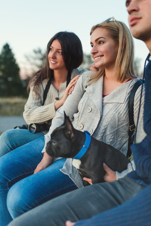 Small group of young people together with black French bulldog puppy siting on wooden bench and looking at sunset. Rear view.の写真素材