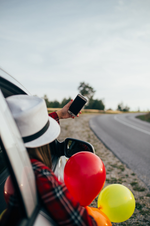 Young woman having fun while holding balloons through car window and taking selfie photo.の写真素材