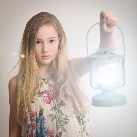 Beautiful young teenage girl with long blond hair holding up an illuminated lantern isolated against a light gray backgroundの写真素材