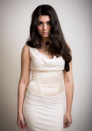 Studio portrait of a beautiful young brunette woman with long hair standing in an elegant white cream dress isolated against a grey backgroundの写真素材