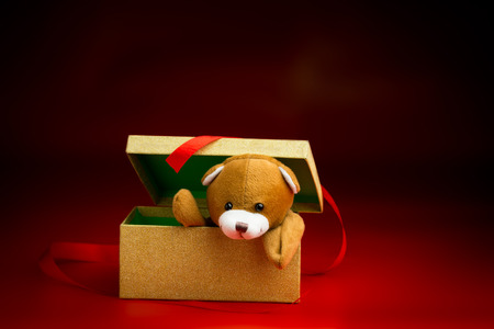 Brown Christmas teddy bear peeking over the edge of a gift box with a loose ribbon isolated against a black and red backgroundの写真素材