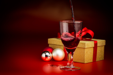 A wine glass is poured with red wine in front of a golden present box with a red ribbon and three baubles next to itの写真素材